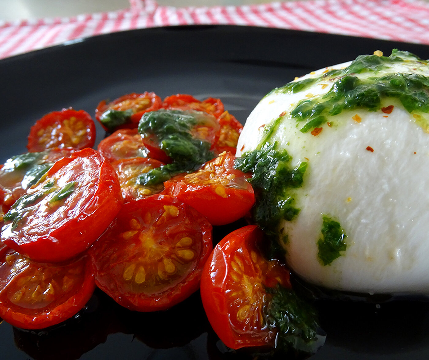The colours of the salad pop against a black plate. White creamy burrata, vibrant red roasted tomatoes, and stark white burrata.
